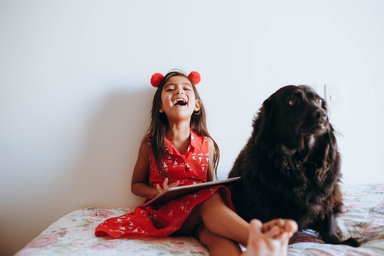 About Joyful child in red dress laughing with a black dog indoors, capturing a heartwarming moment.